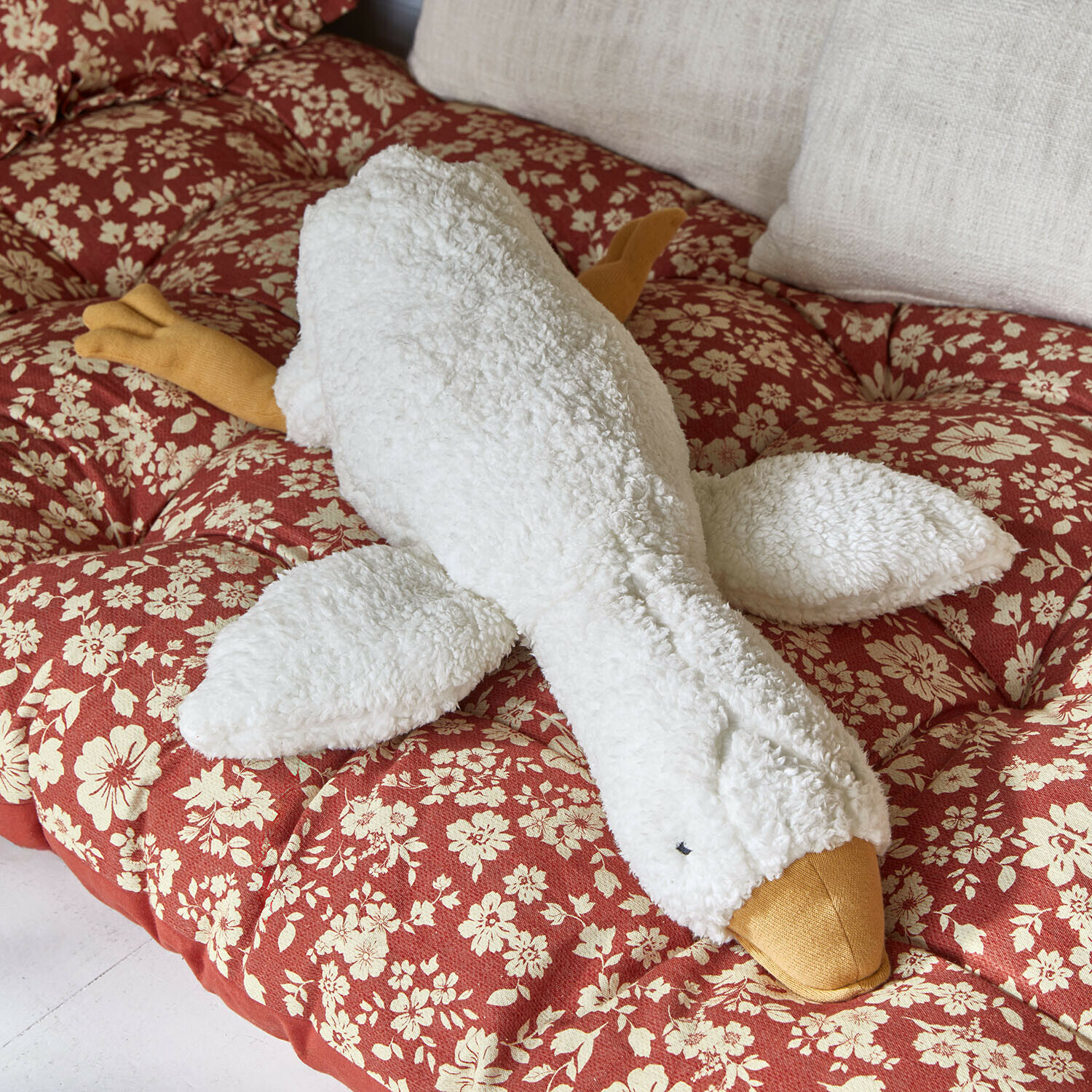 Peluche Tillyboo Peluche oie blanche et jaune posée sur un coussin rouge à motif floral pour décoration intérieure