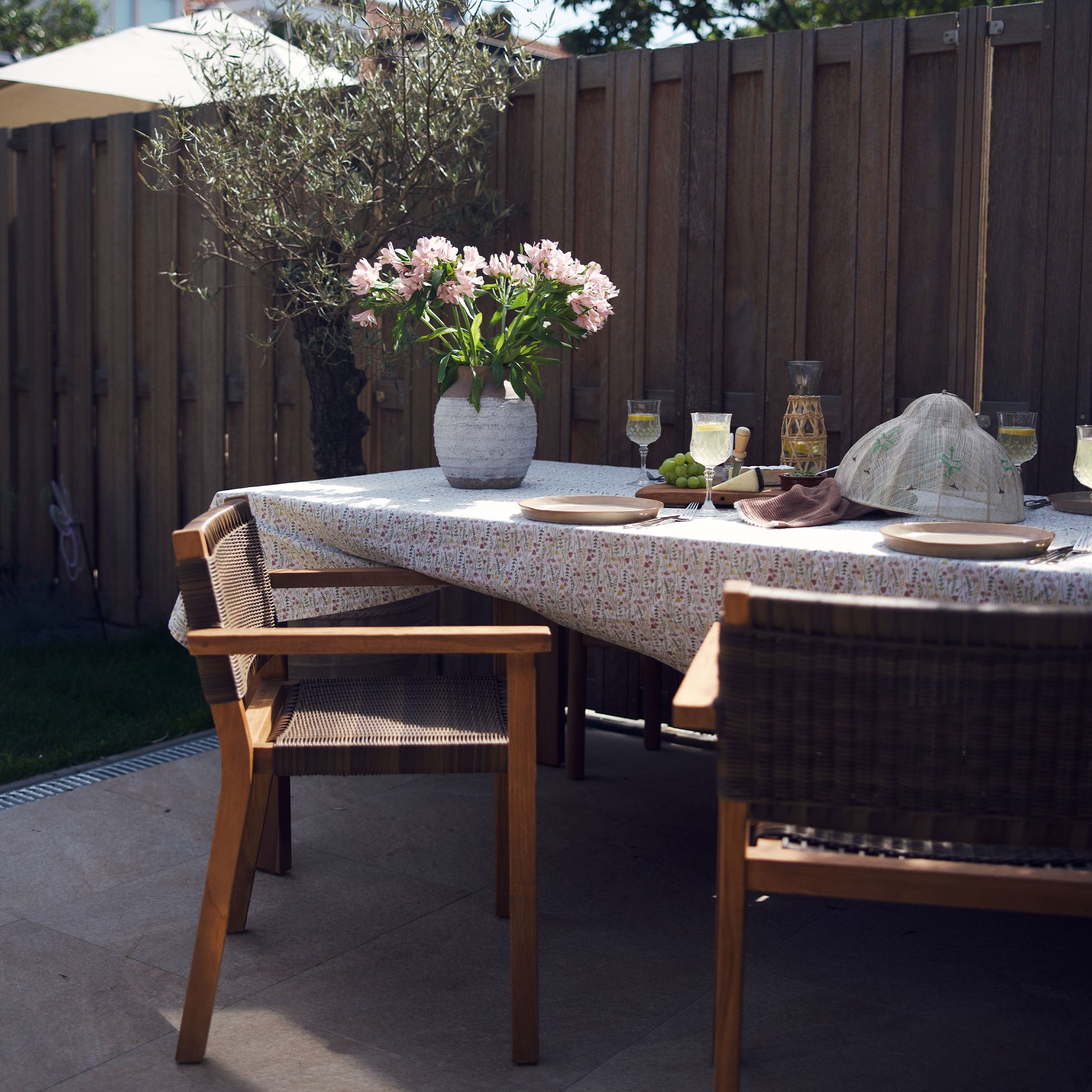 Table de jardin en bois avec chaises, vase en pierre avec fleurs roses sur nappe à motif floral