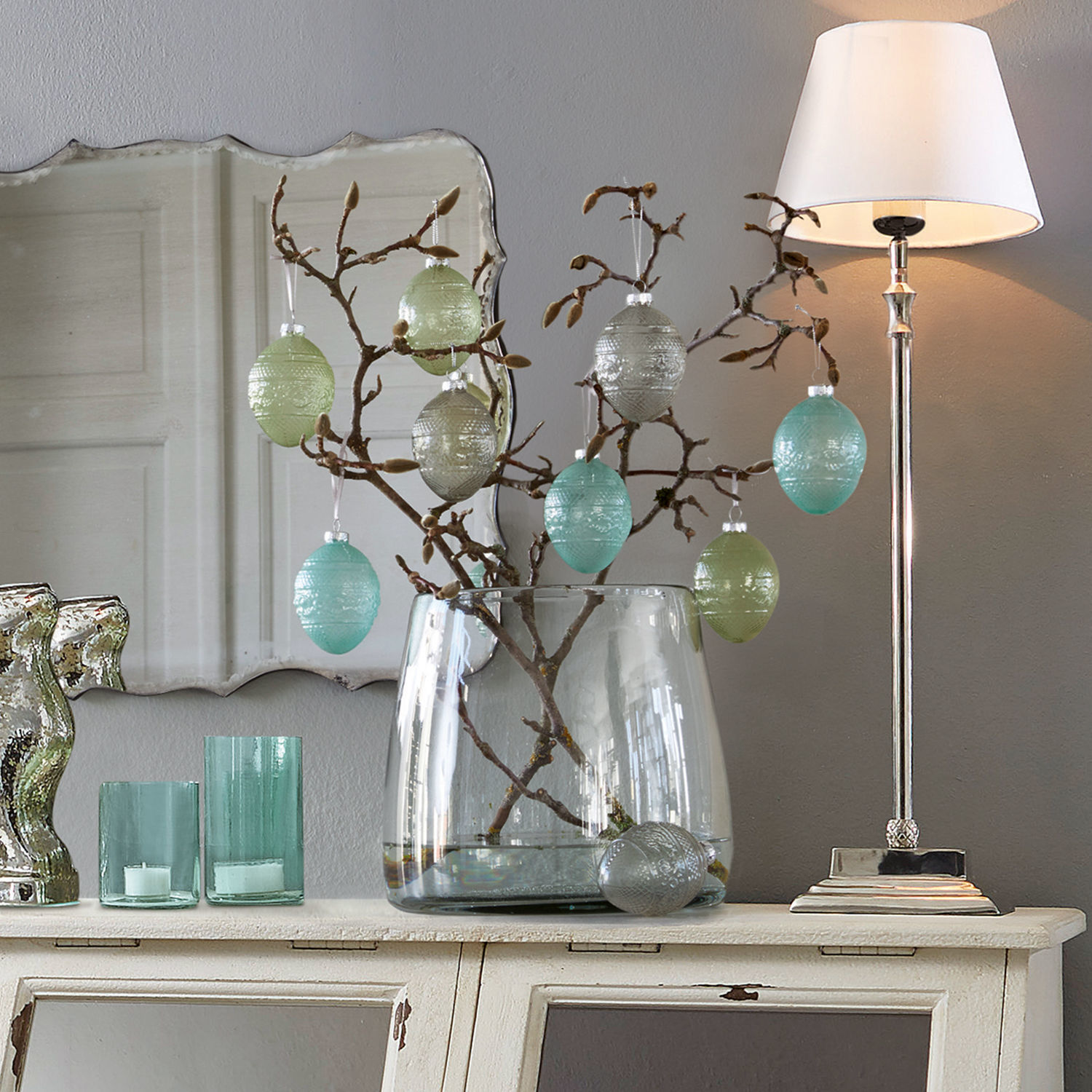 Vase Inga Oeufs en verre décoratifs pastel suspendus sur une branche dans un vase transparent sur une commode blanche