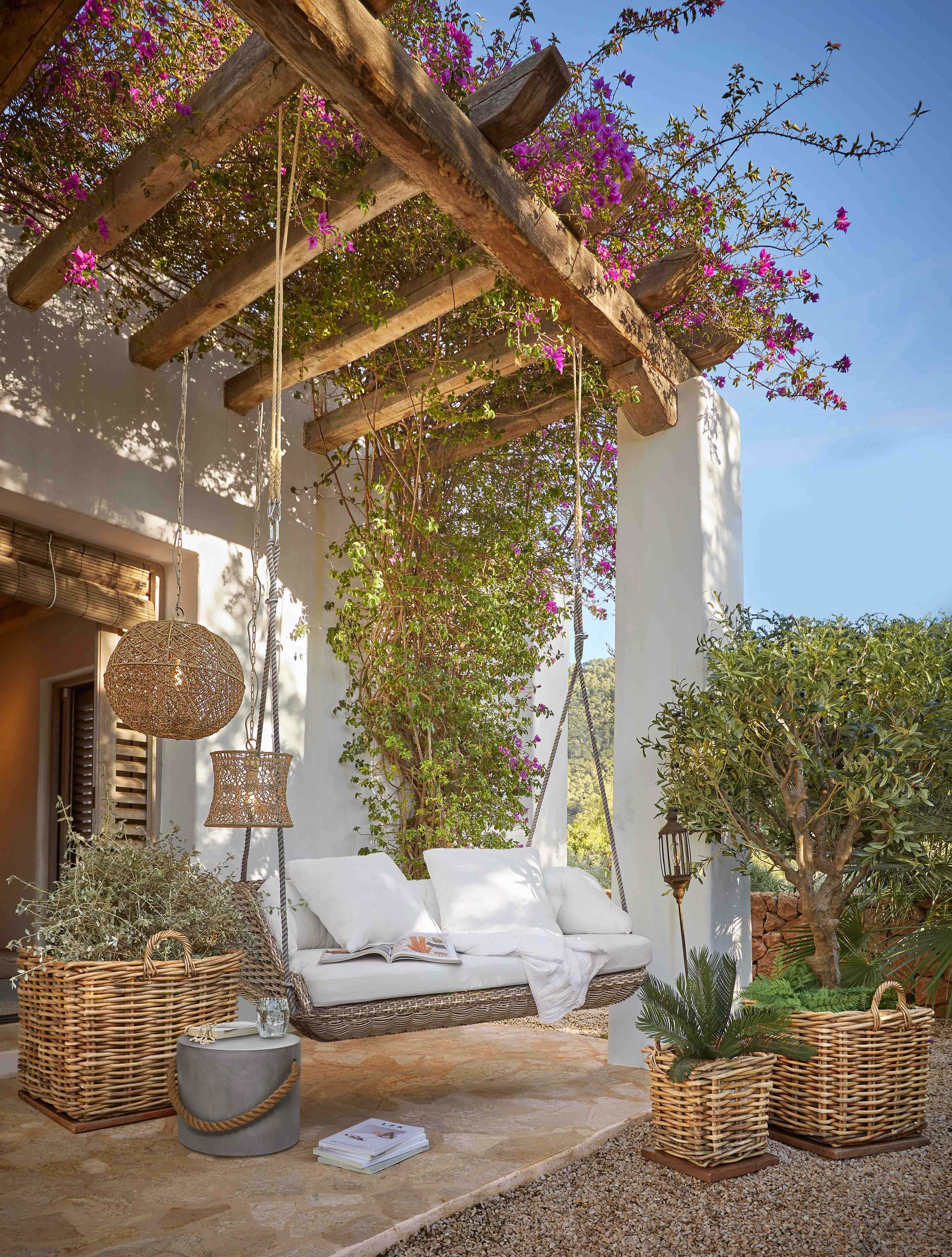 Balançoire en osier avec coussins blancs sous une pergola en bois ornée de fleurs roses éclatantes