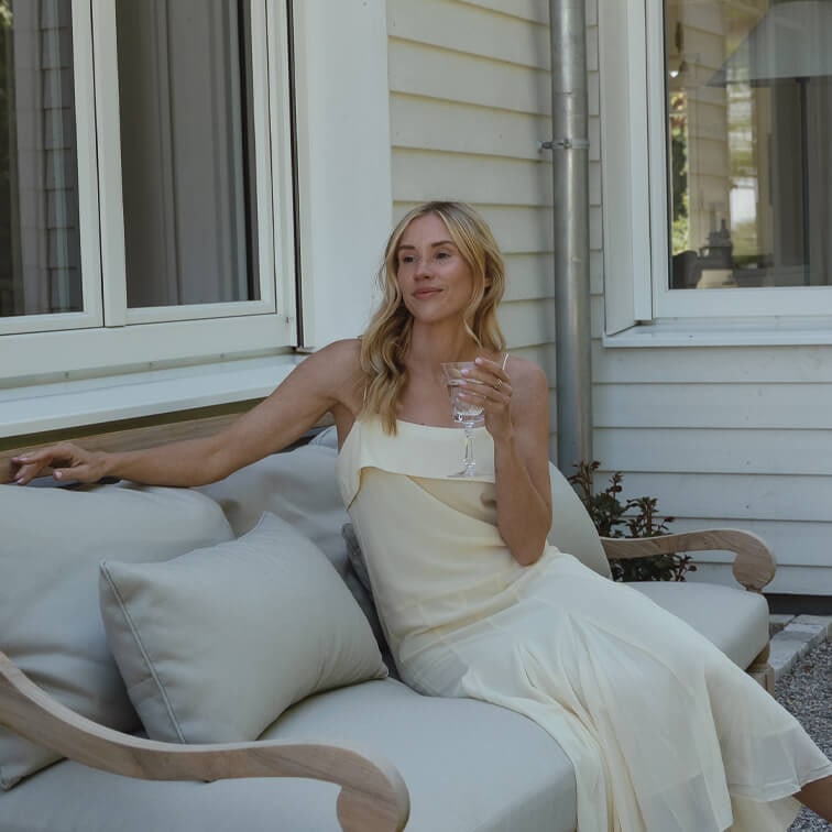 Femme en robe crème assise sur un canapé gris clair à l'extérieur, tenant un verre à vin, détente sur la terrasse