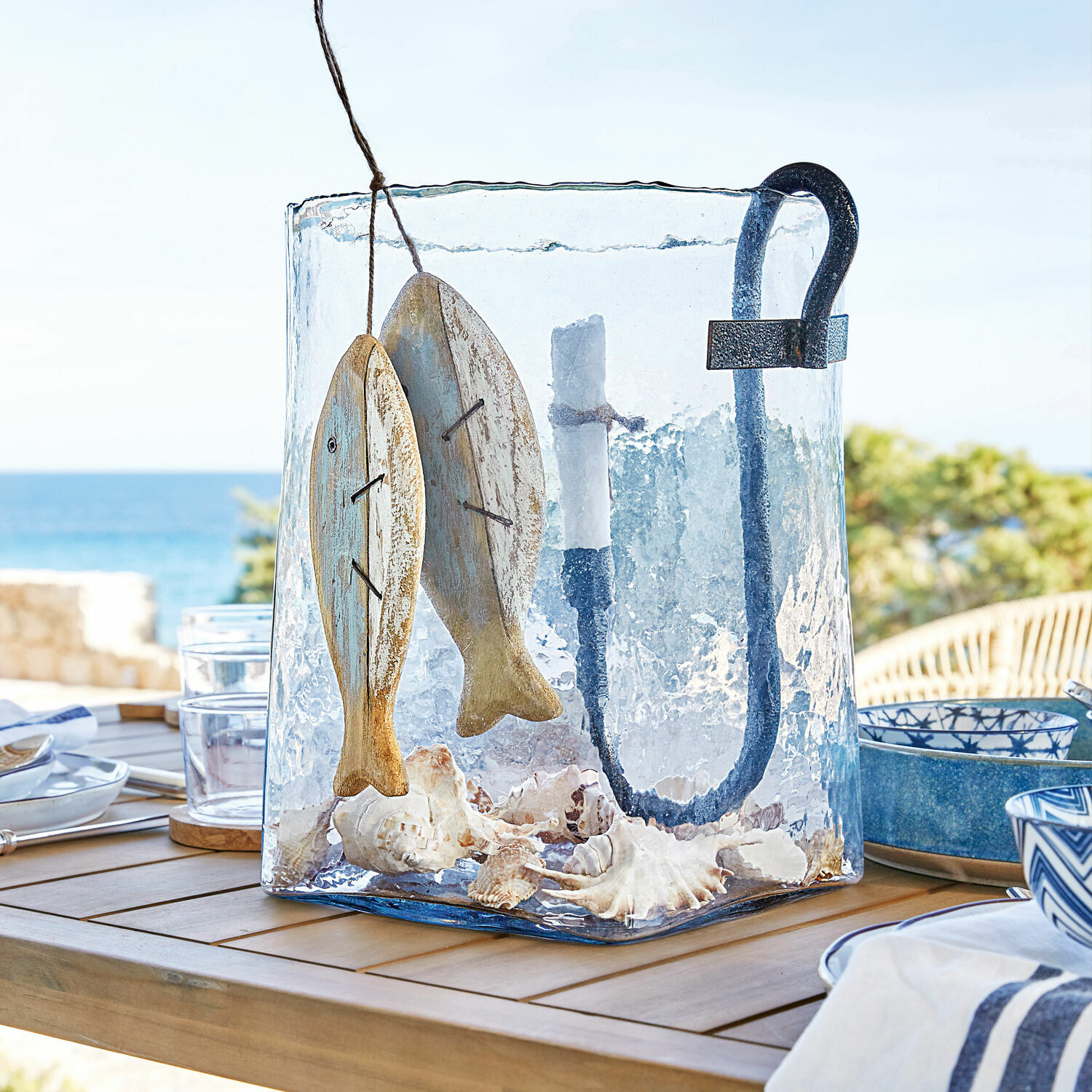 Photophore Arcene Lanterne en verre avec poignée en métal, décor poissons en bois et coquillages sur table en bois en terrasse