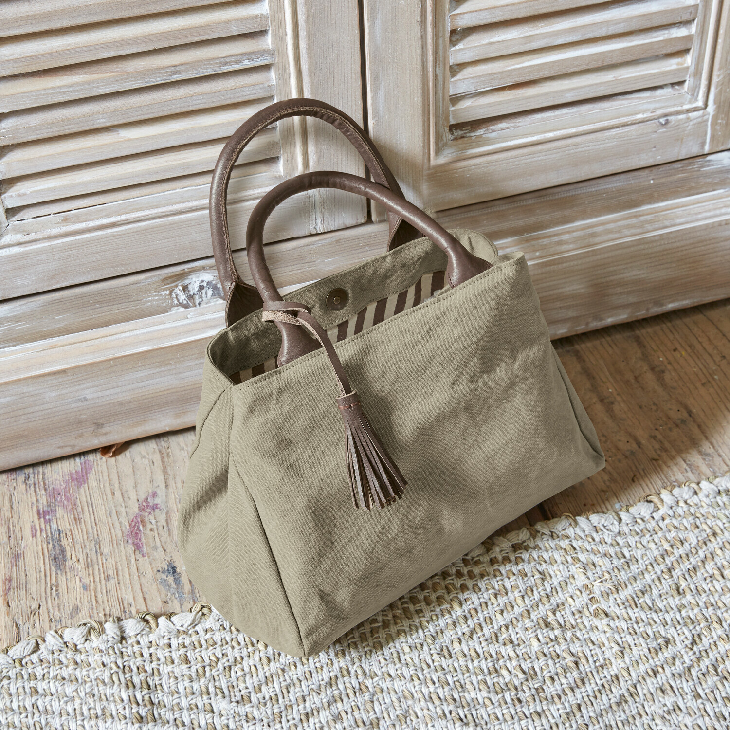 Sac Hoya Sac beige en tissu avec poignées en cuir marron et pompon posé sur un sol en bois