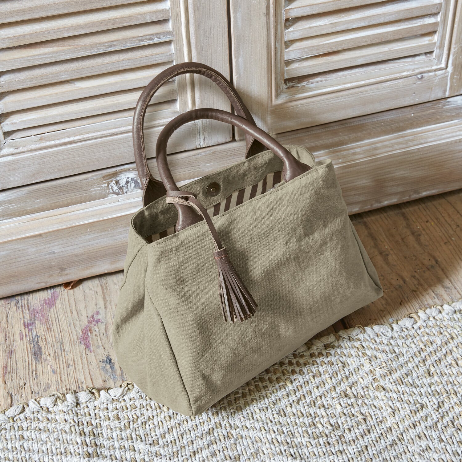 Sac Hoya Sac beige en tissu avec poignées en cuir marron et pompon posé sur un sol en bois