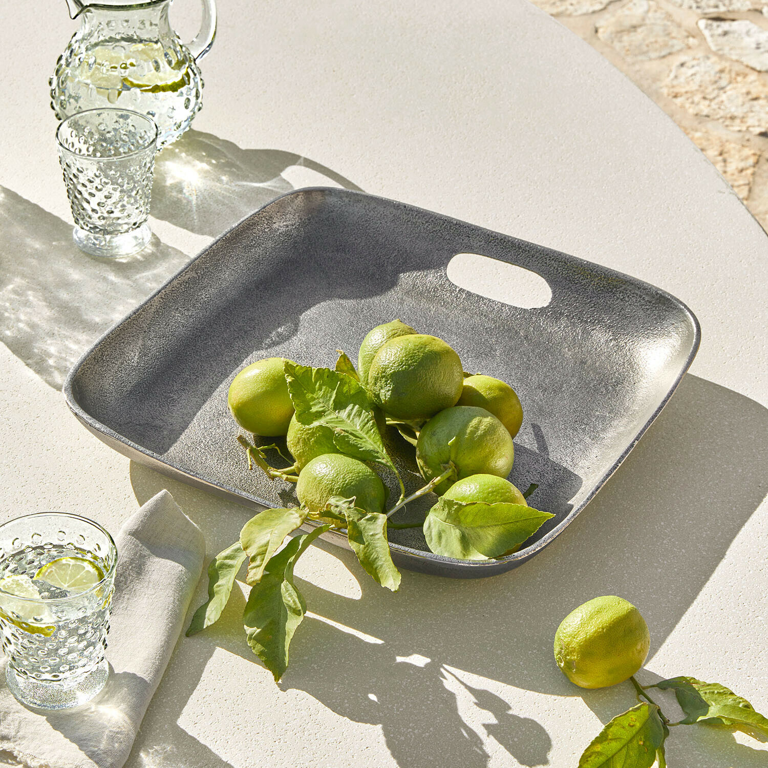 Plateau carré en métal gris avec citrons verts sur une table claire, décoration de table estivale