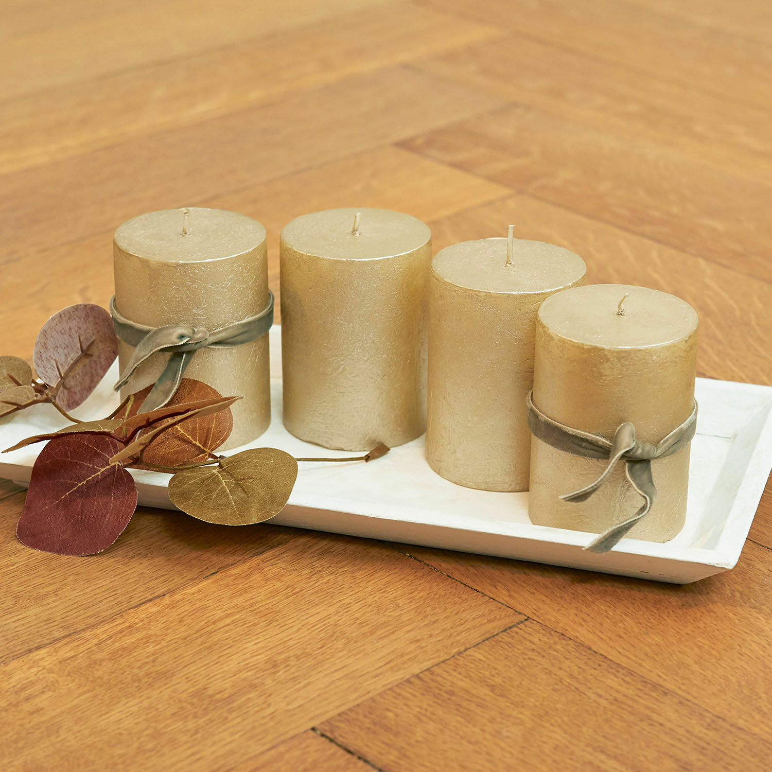 Lot de 4 bougies Virrans Ensemble de quatre bougies décoratives dorées sur un plateau blanc avec feuilles décoratives sur table en bois
