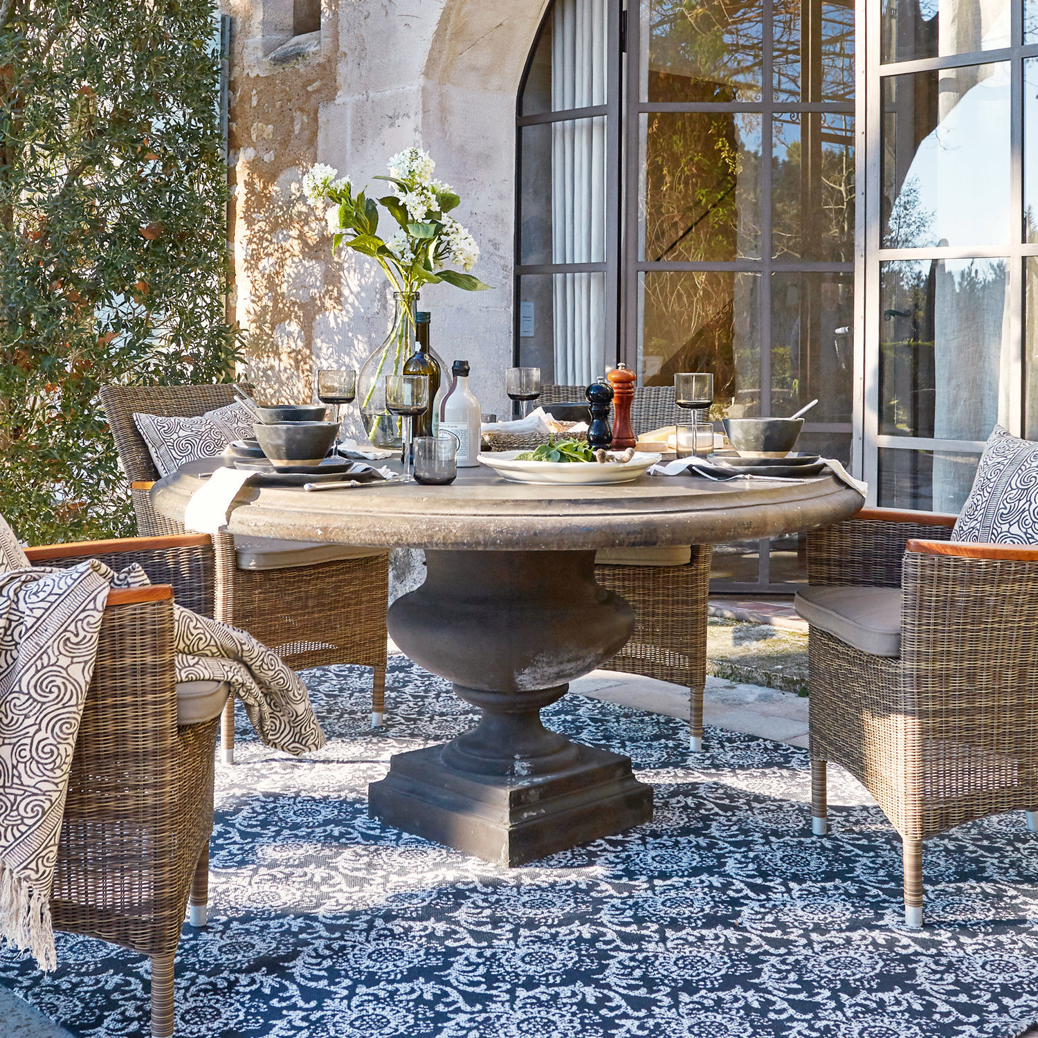 Table Monterey Table ronde en pierre avec base noire entourée de chaises en osier sur terrasse avec tapis décoratif