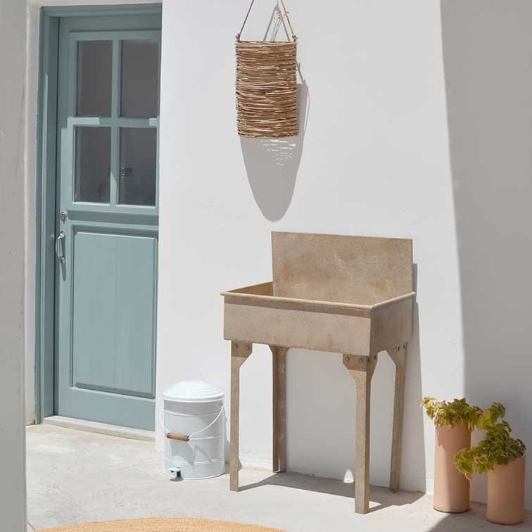 Jardinière beige en pierre sur pieds devant un mur blanc avec porte bleue et panier en osier suspendu