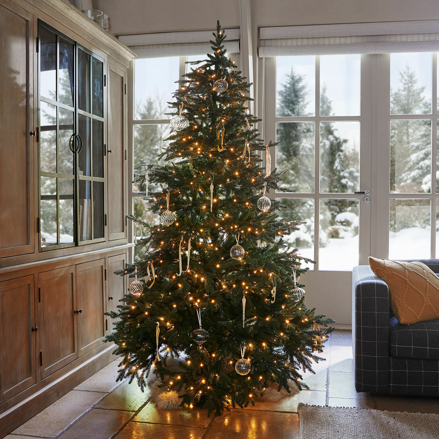 Sapin de Noël Celindor Sapin de Noël illuminé avec branches vertes et décorations en verre dans un salon chaleureux avec vue sur la neige