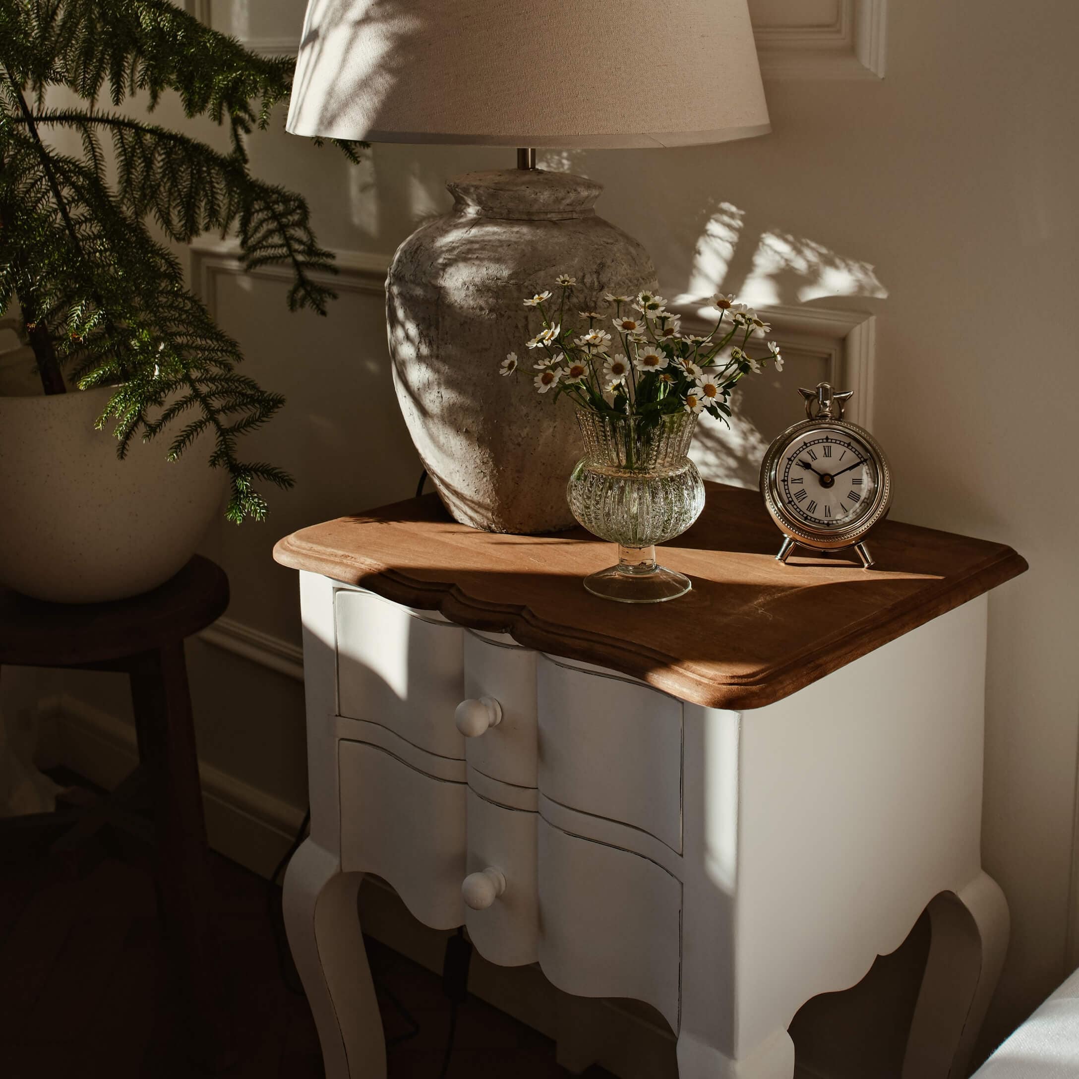 Table de chevet en bois blanc avec plateau marron, lampe, vase en verre avec fleurs et horloge argentée