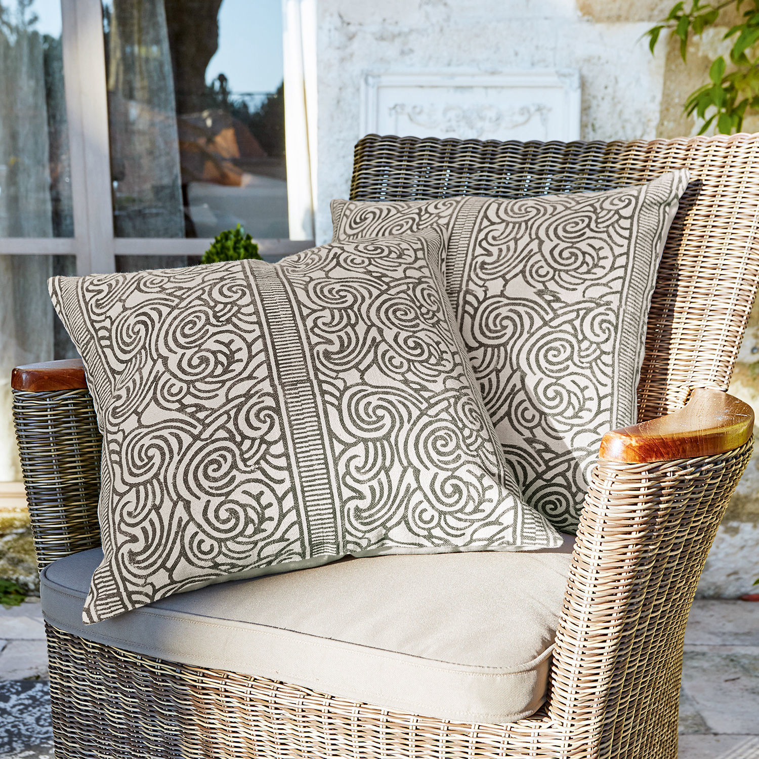 Lot de 2 housses de coussins Ensis Deux coussins décoratifs en coton beige avec motif marron sur fauteuil en osier sur terrasse