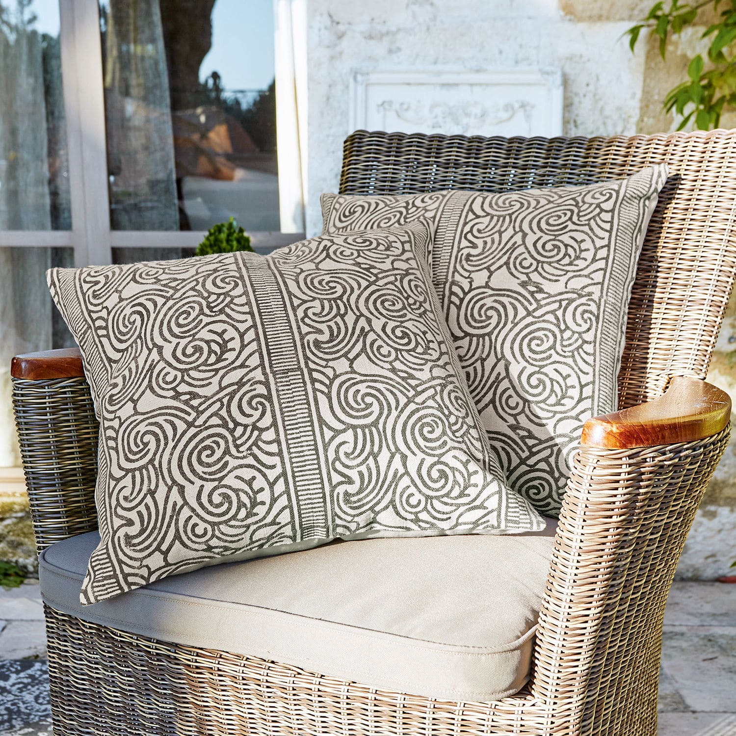 Lot de 2 housses de coussins Ensis Deux coussins décoratifs en coton beige avec motif marron sur fauteuil en osier sur terrasse