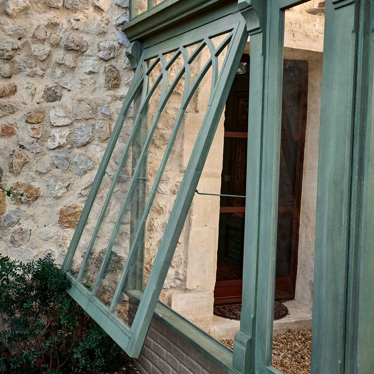 Fenêtre verte en métal avec motif décoratif sur mur en pierre et plantes en arrière-plan