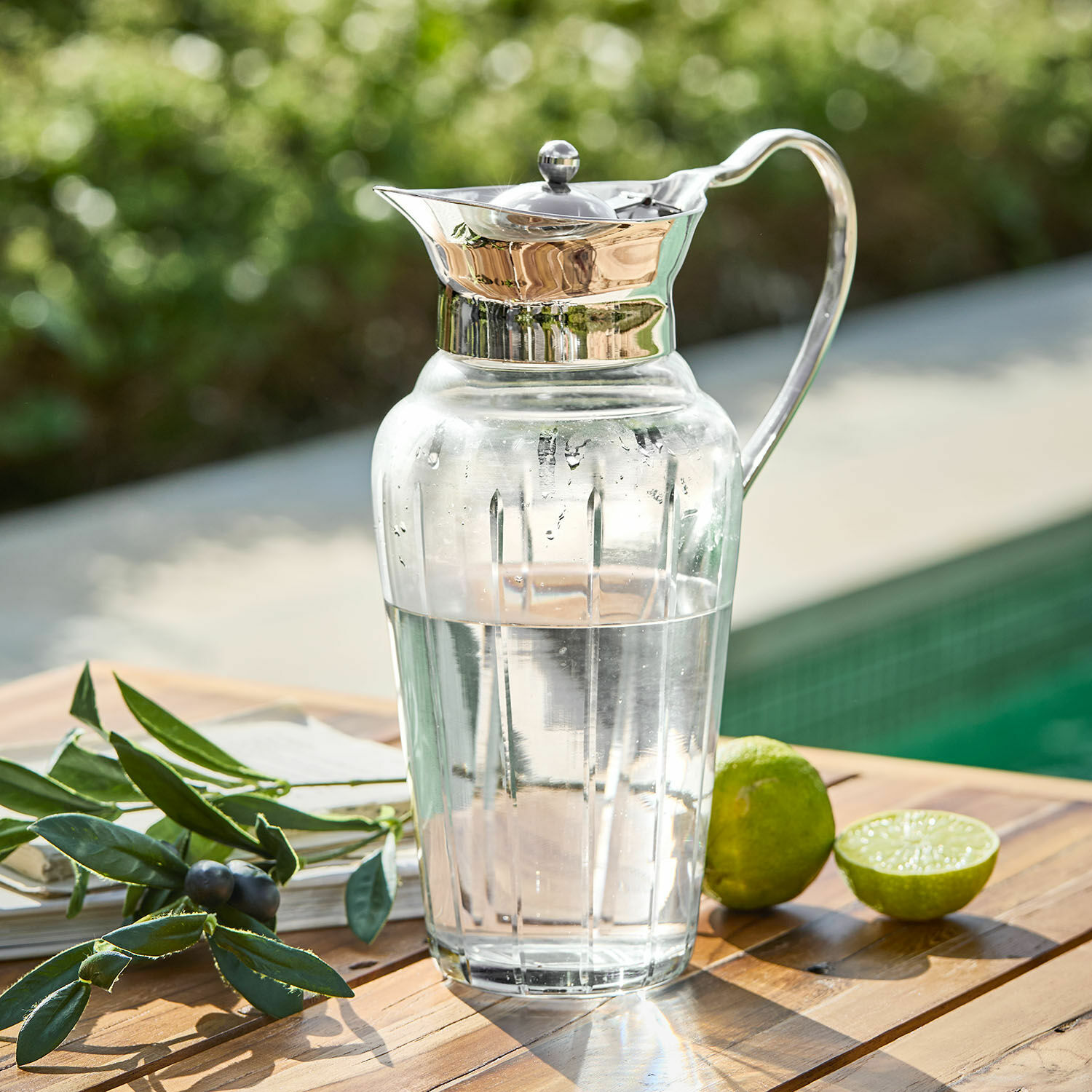 Pichet Braux Carafe en verre avec couvercle et poignée en métal remplie d'eau, posée sur une table en bois près d'un citron vert et d'une branche