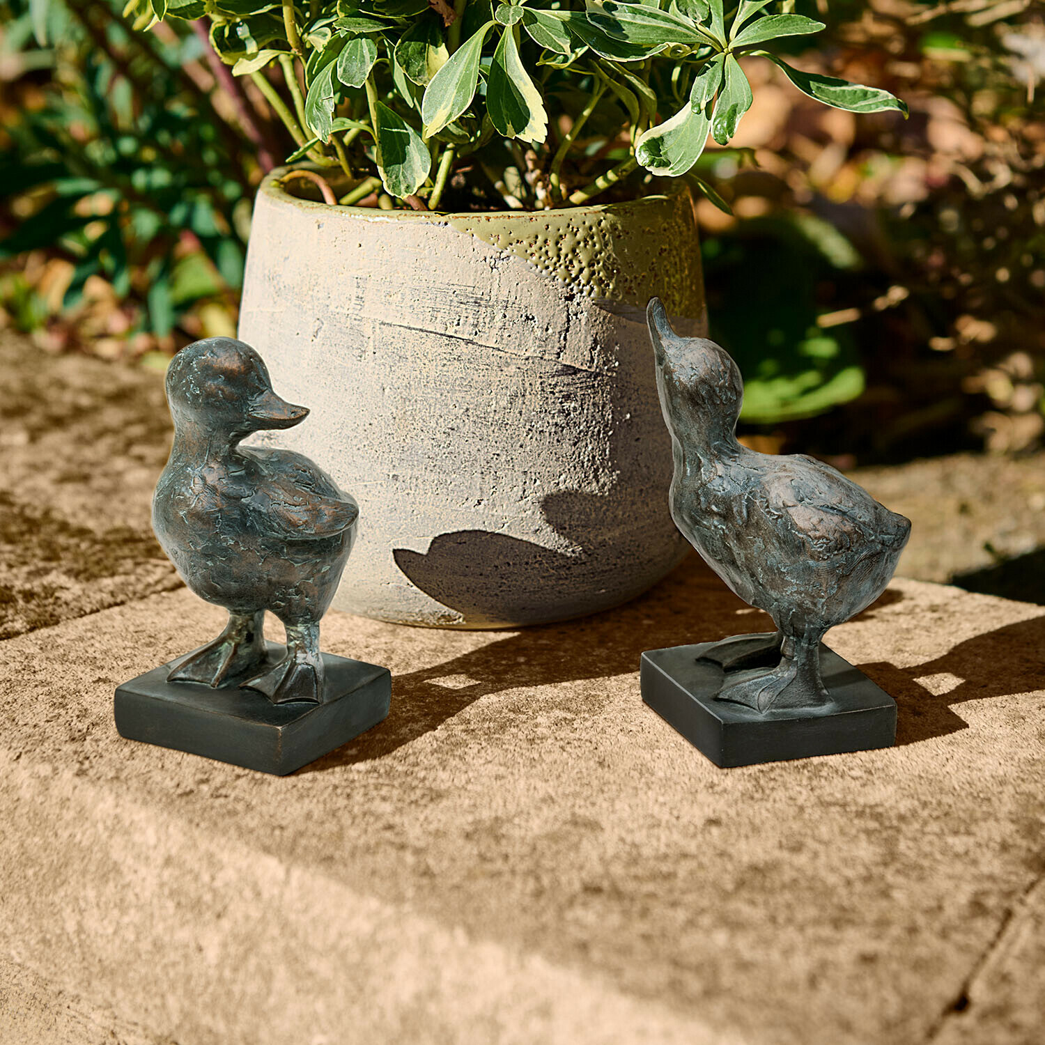 Deux figurines de canards en métal foncé posées sur une surface en pierre près d'un pot de fleurs