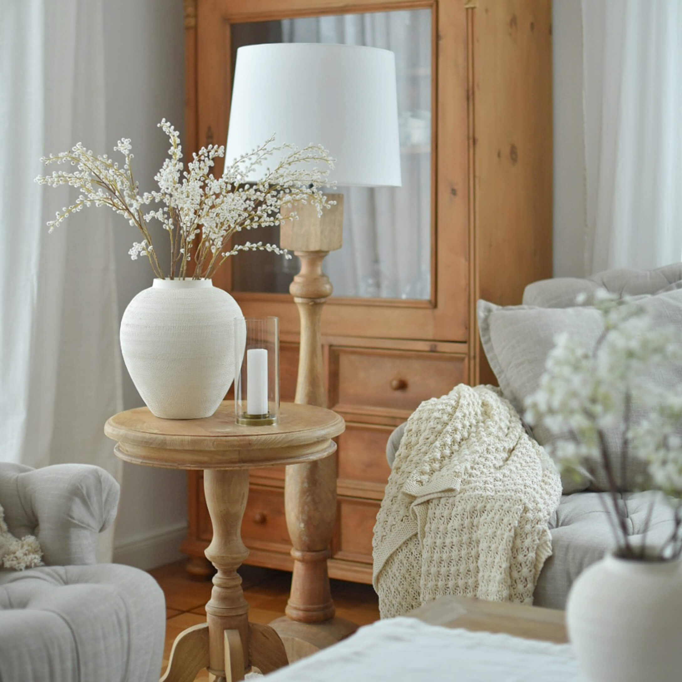 Table en bois clair avec vase en céramique blanche et lampe dans un salon cosy avec plaid beige
