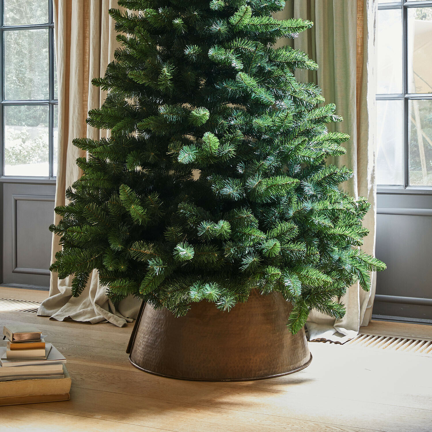 Bordure de l"arbre de Noël Omeyra Sapin artificiel dans un cache-pot en métal cuivré posé sur un sol en bois clair près de livres