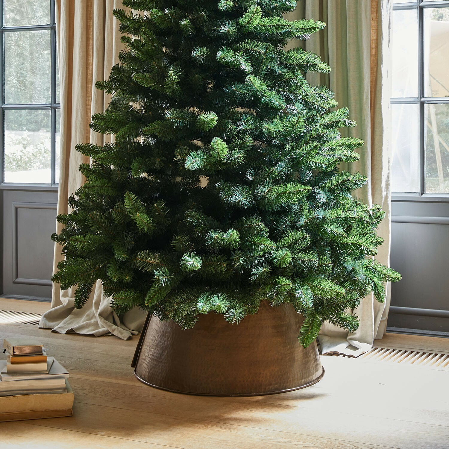 Bordure de l"arbre de Noël Omeyra Sapin artificiel dans un cache-pot en métal cuivré posé sur un sol en bois clair près de livres