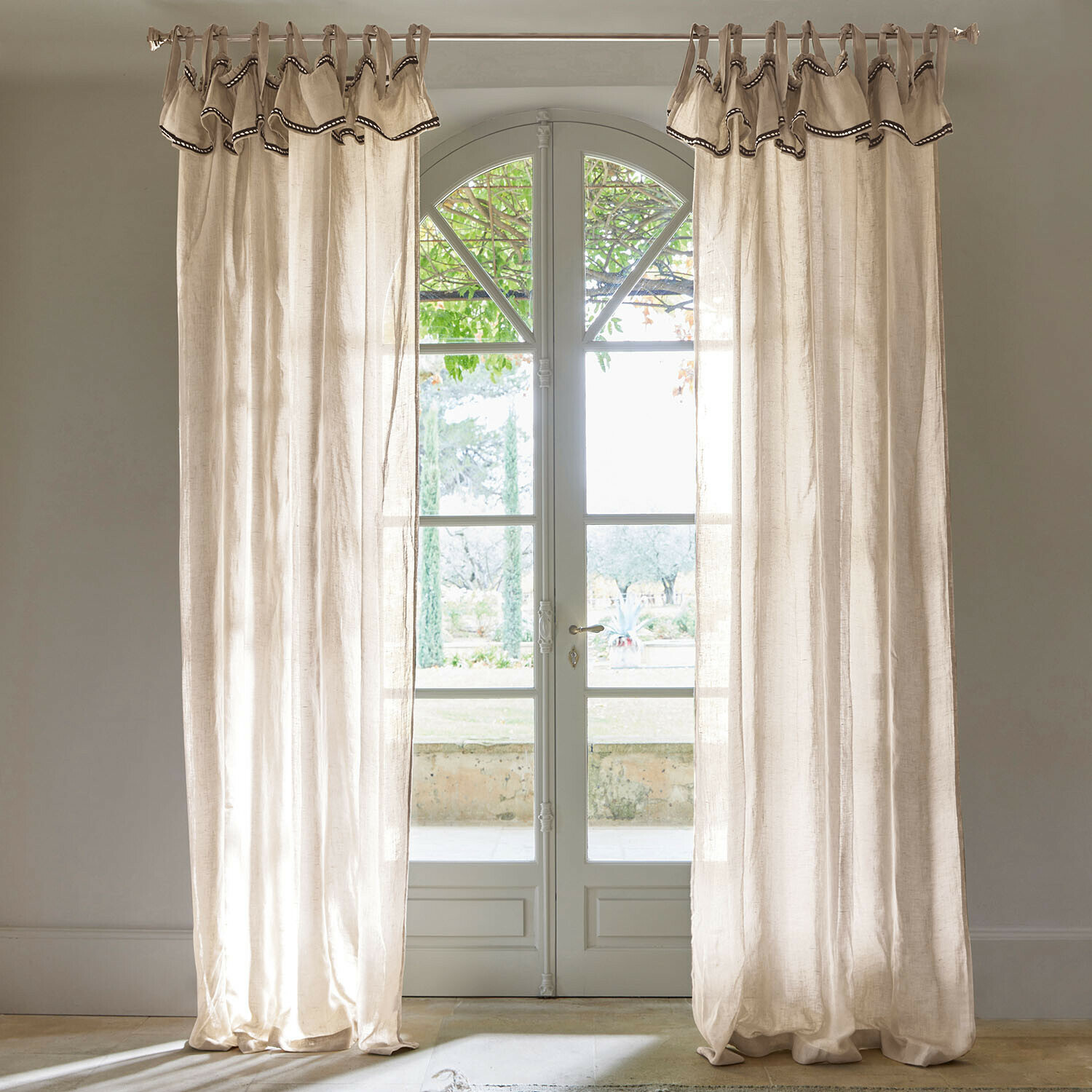 Rideau Romagny Rideaux en lin beige avec bordure décorative suspendus devant une porte-fenêtre donnant sur un jardin