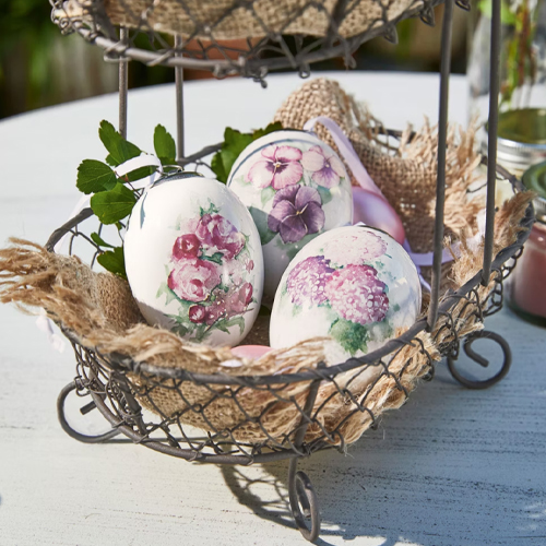 Trois œufs en céramique blanche avec motifs floraux dans un panier en métal avec toile de jute sur une table
