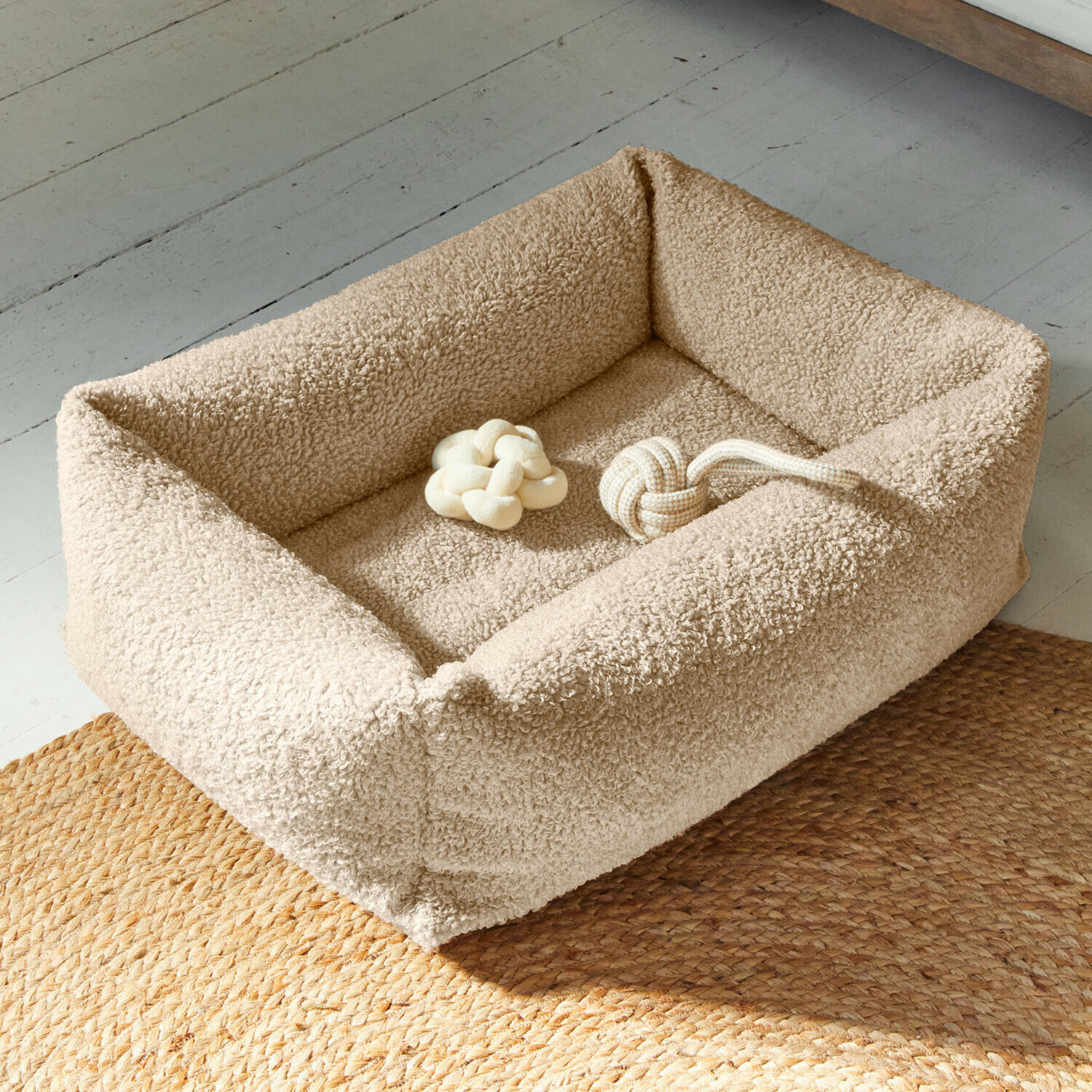Lit pour animaux Giolena Panier pour chien beige en tissu doux avec deux jouets nœuds beiges sur un sol avec tapis tressé