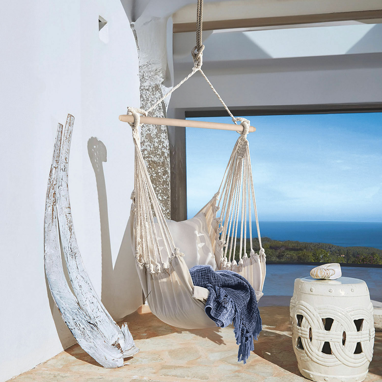 Fauteuil suspendu Leano Hamac beige en coton avec barre en bois et couverture bleue sur terrasse avec vue sur la mer
