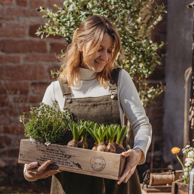 Femme en salopette en velours côtelé marron tenant une caisse en bois avec des plantes vertes dans un jardin