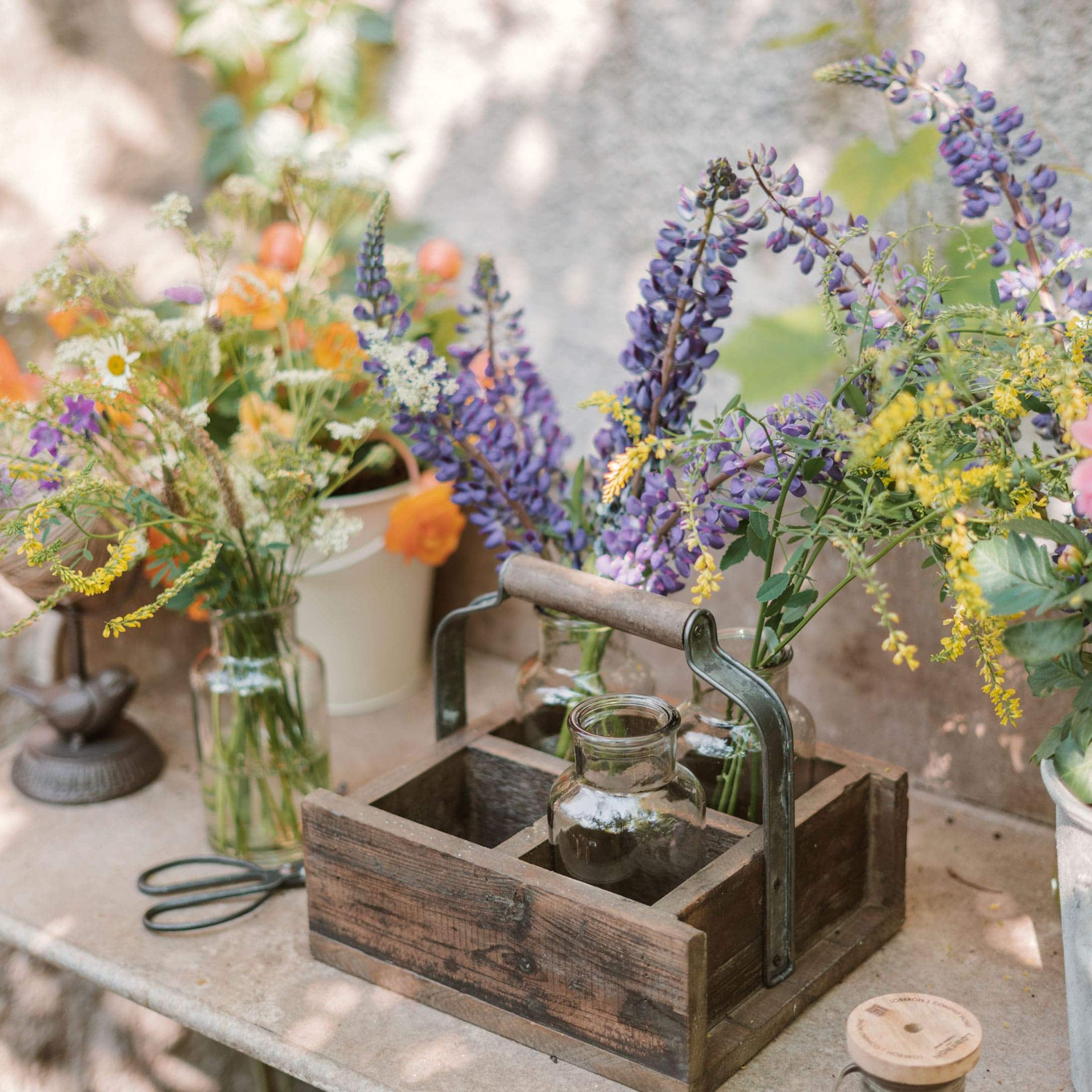 Organisateur en bois avec poignée et vases en verre contenant des fleurs colorées sur une table extérieure