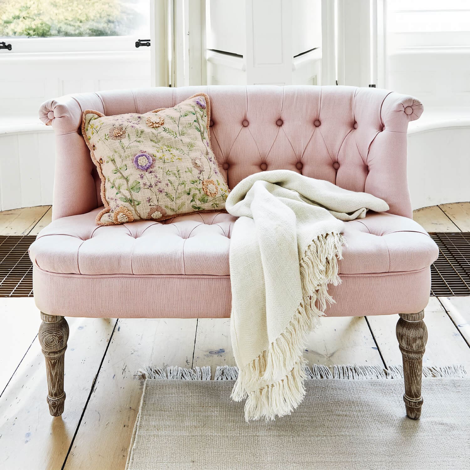 Canapé rose rembourré avec pieds en bois, coussin décoratif et plaid crème sur un sol clair
