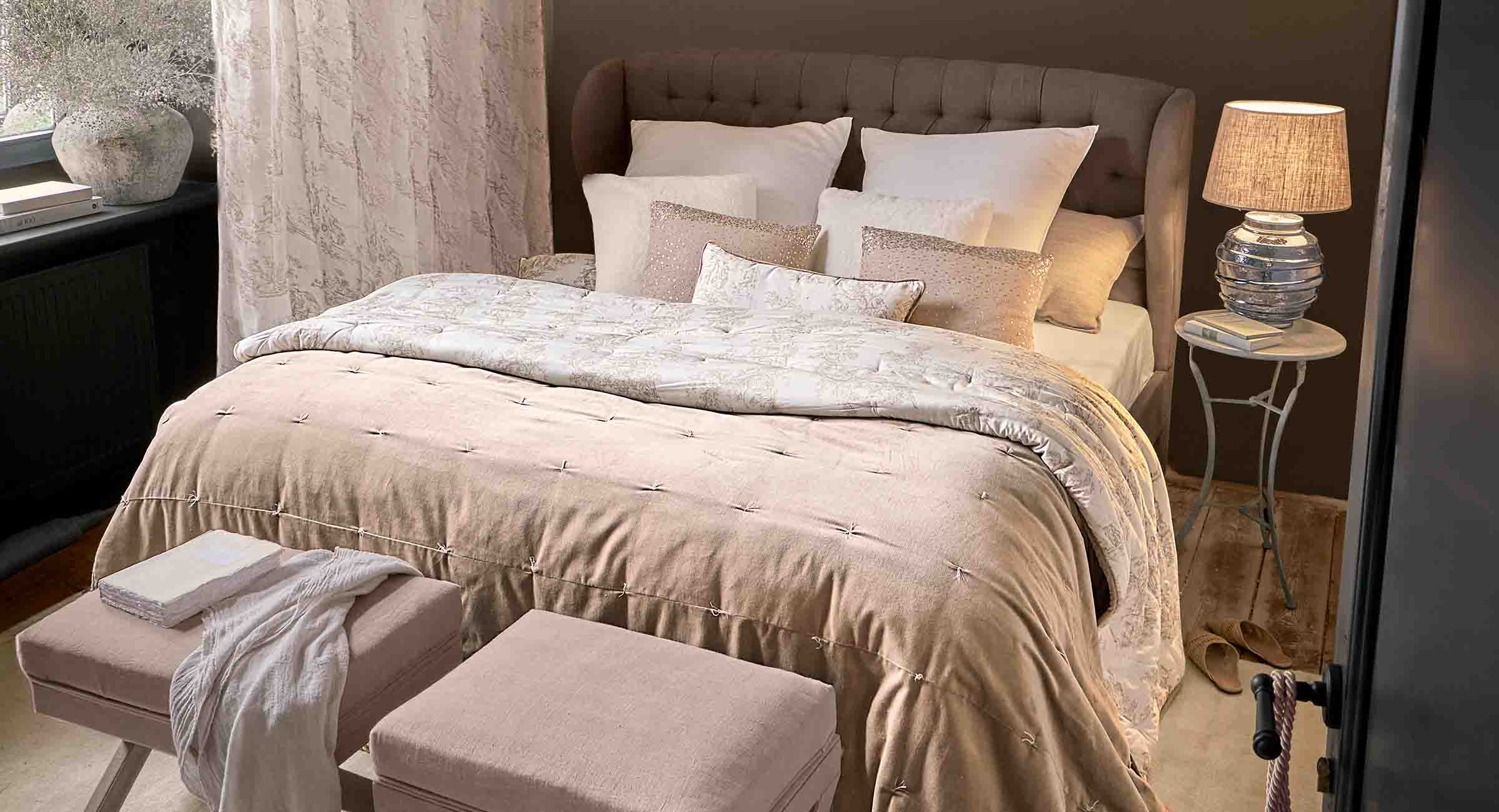 Chambre chaleureuse avec couette beige et coussins sur lit capitonné, lampe posée sur table de chevet