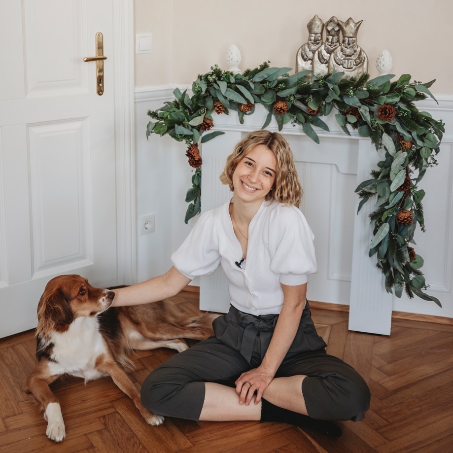 Jeune femme en pull blanc et pantalon sombre assise sur un sol en bois avec un chien près d'une décoration de Noël en feuillage vert et pommes de pin
