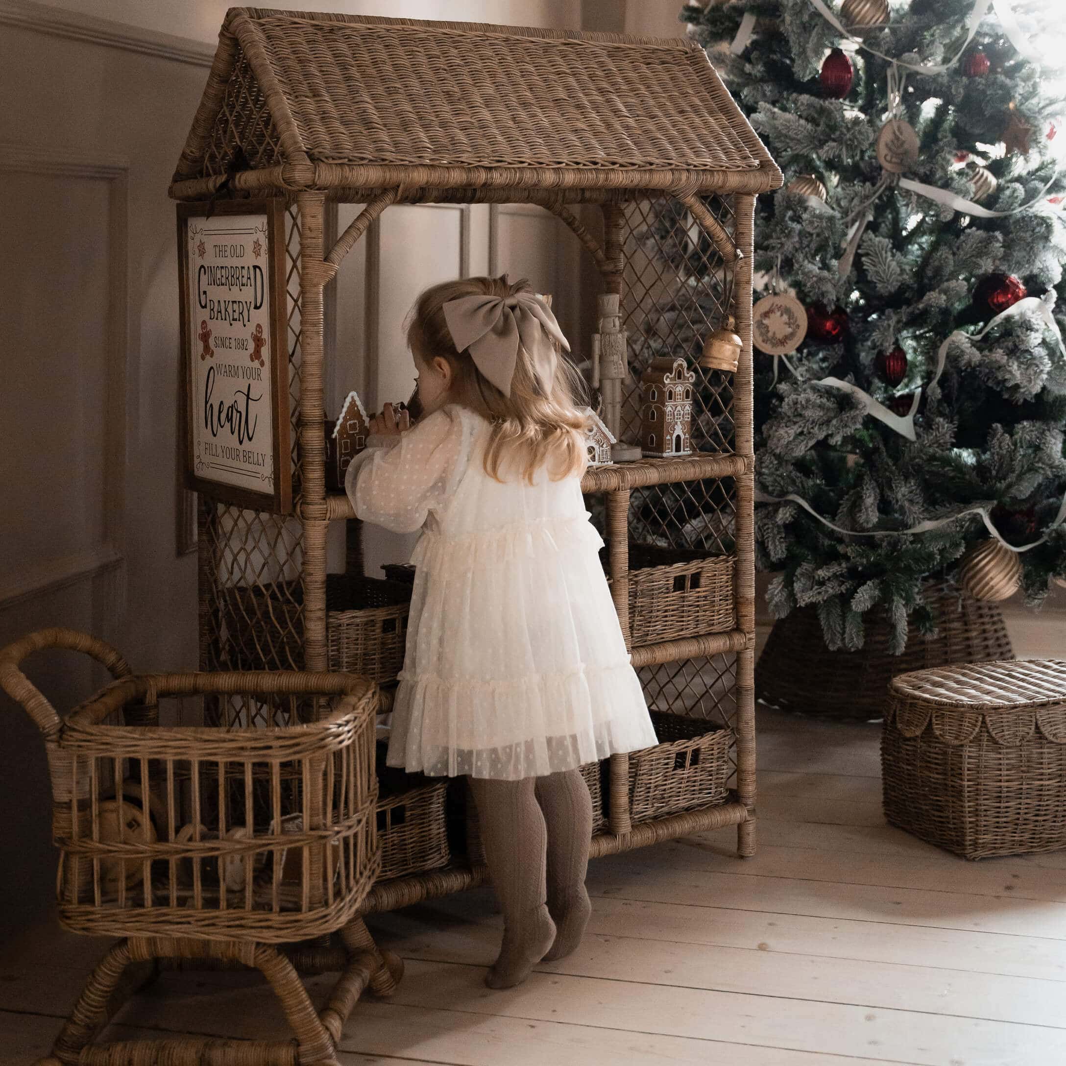 Enfant en robe blanche devant une étagère en osier avec des maisons en pain d'épice près d'un sapin de Noël décoré