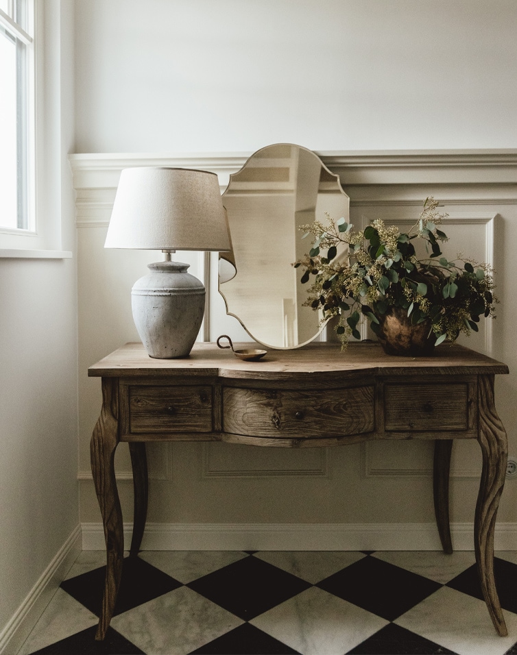 Bureau en bois avec miroir, lampe et plante décorative sur sol noir et blanc dans une pièce lumineuse
