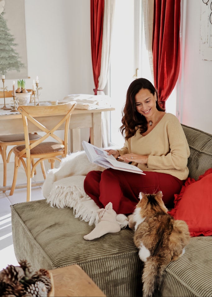 Femme en pull beige et pantalon rouge lisant un livre sur un canapé vert avec un chat dans un intérieur chaleureux.