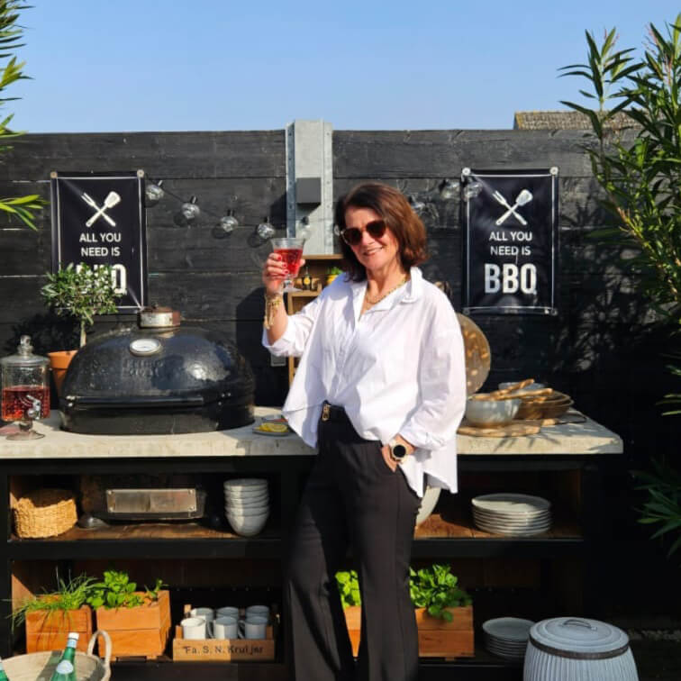 Femme en chemise blanche et pantalon noir près d'un barbecue extérieur avec panneau BBQ sur fond de mur noir