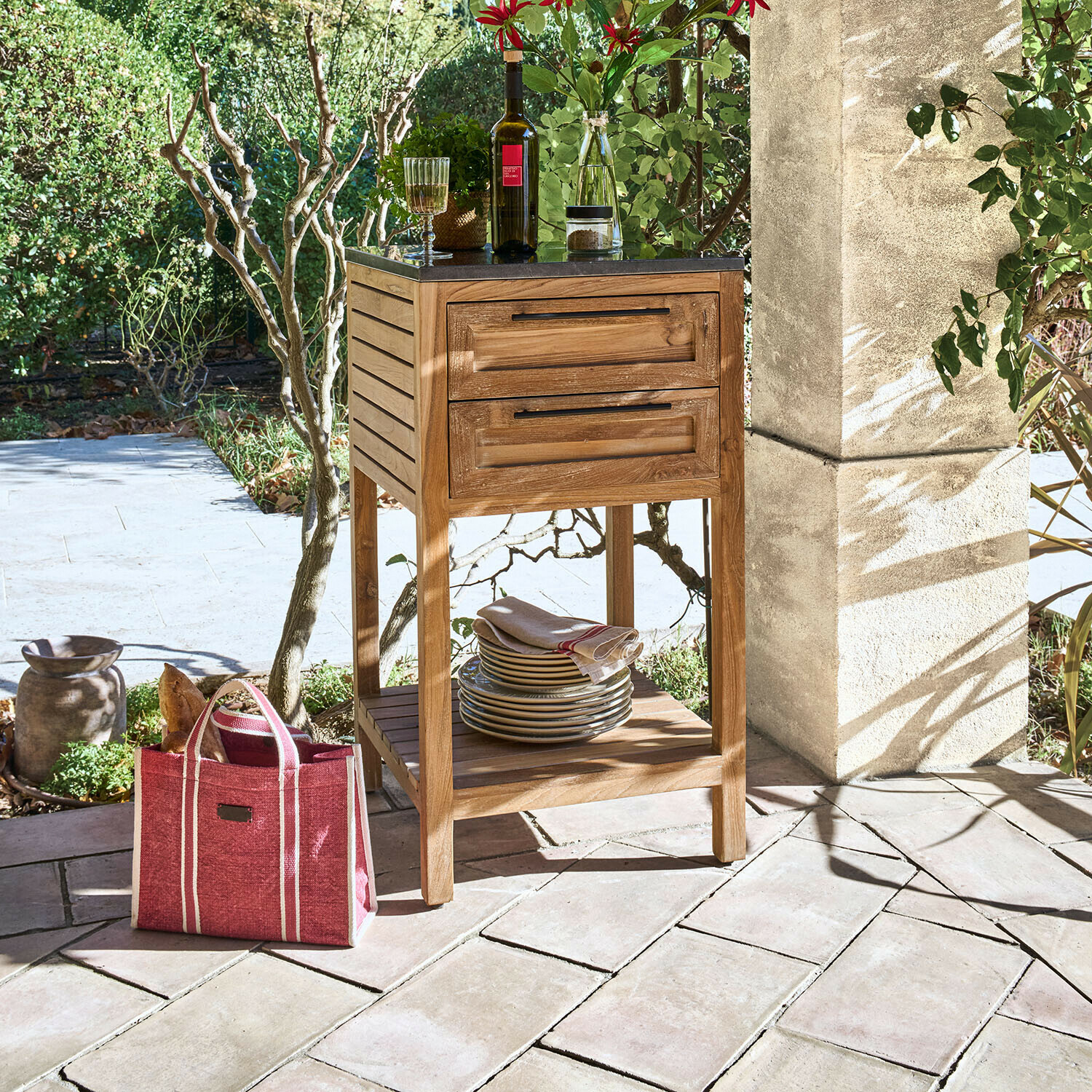 Meuble de cuisine d’extérieur Corviano Meuble en bois avec plateau noir et deux tiroirs sur terrasse avec vaisselle et bouteille de vin