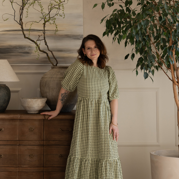 Femme en robe à carreaux verts posant près d'une commode en bois dans un intérieur chaleureux avec plantes