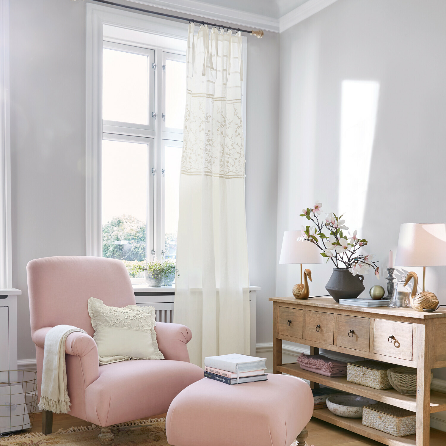 Rideau Lunérie Fauteuil rose avec repose-pieds et coussin crème dans un salon lumineux avec commode en bois et décorations