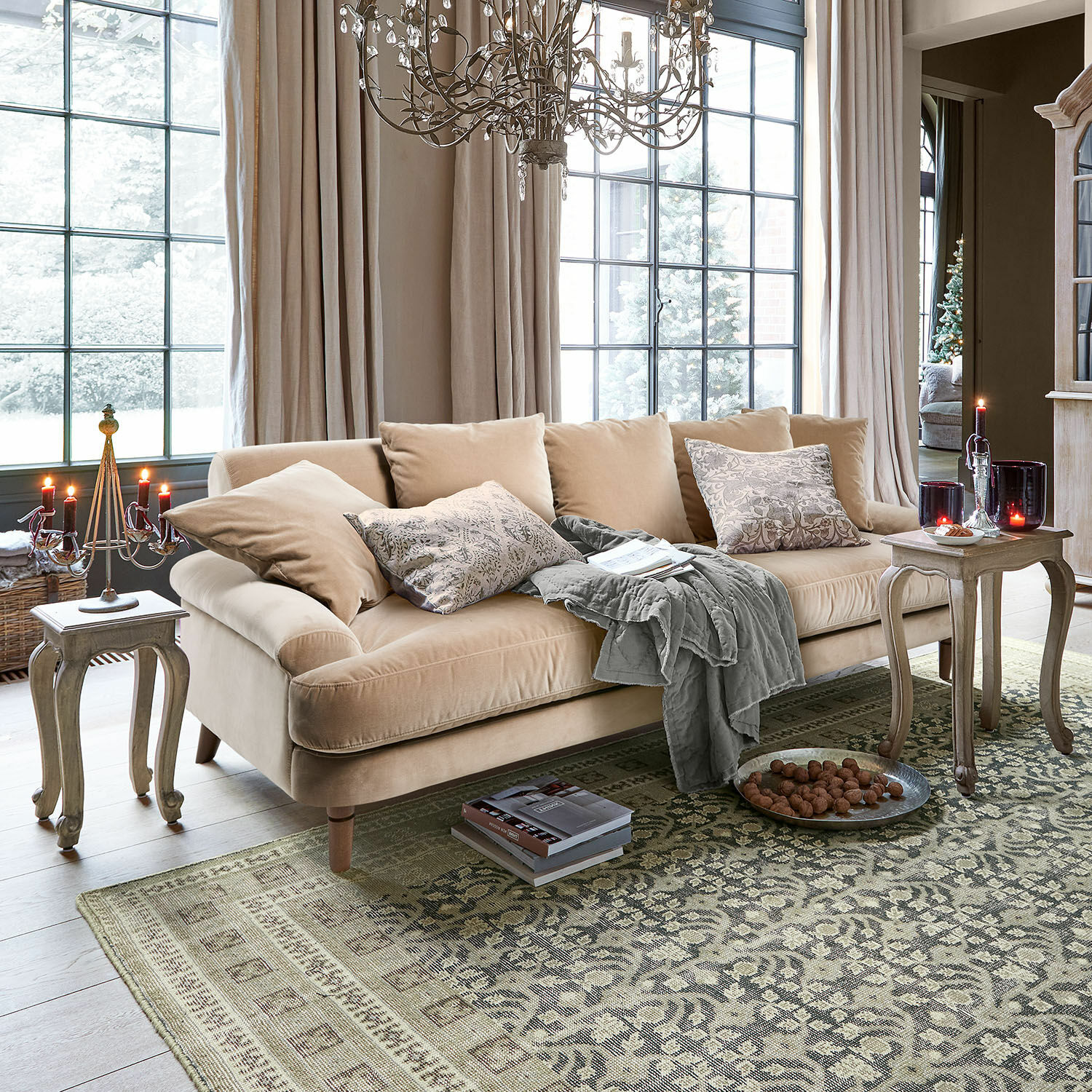 Canapé Rivermere Canapé beige en velours avec coussins et plaid gris dans un salon lumineux avec tapis et bougies