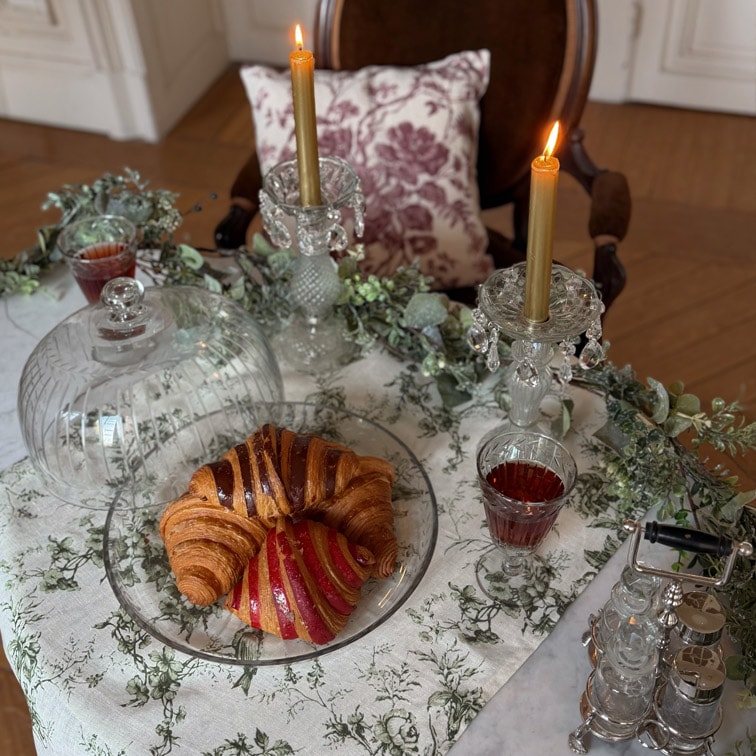 Assiette en verre avec croissant et pâtisserie sur table avec nappe blanche, deux bougies dorées dans des chandeliers en cristal
