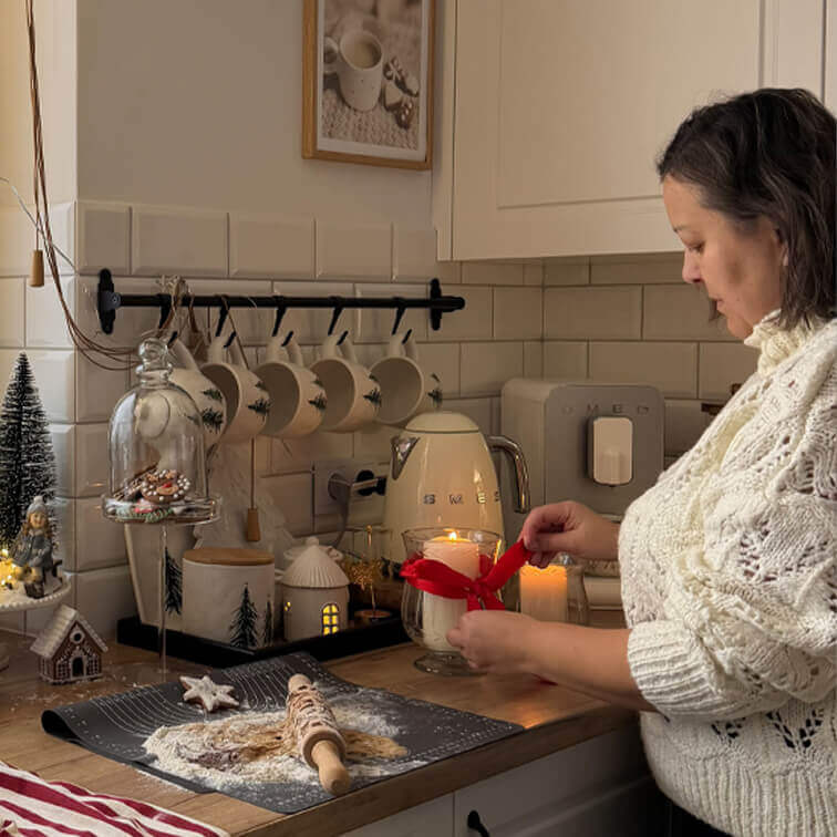 Femme en pull blanc décorant une bougie avec un ruban rouge dans une cuisine avec plan de travail en bois et décorations de Noël