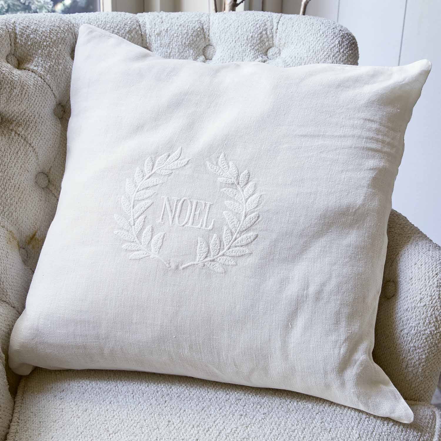 Coussin blanc en lin avec broderie NOEL et couronne décorative sur fauteuil clair, accessoire de Noël
