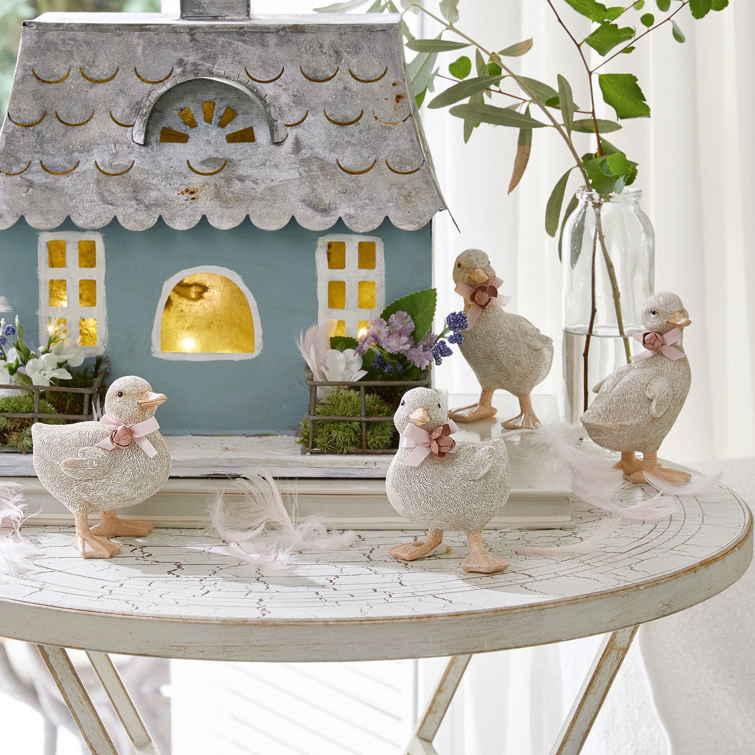 Lot de 4 canetons Orélie Figurines décoratives de canards en matériau clair sur une table blanche avec maison et fleurs
