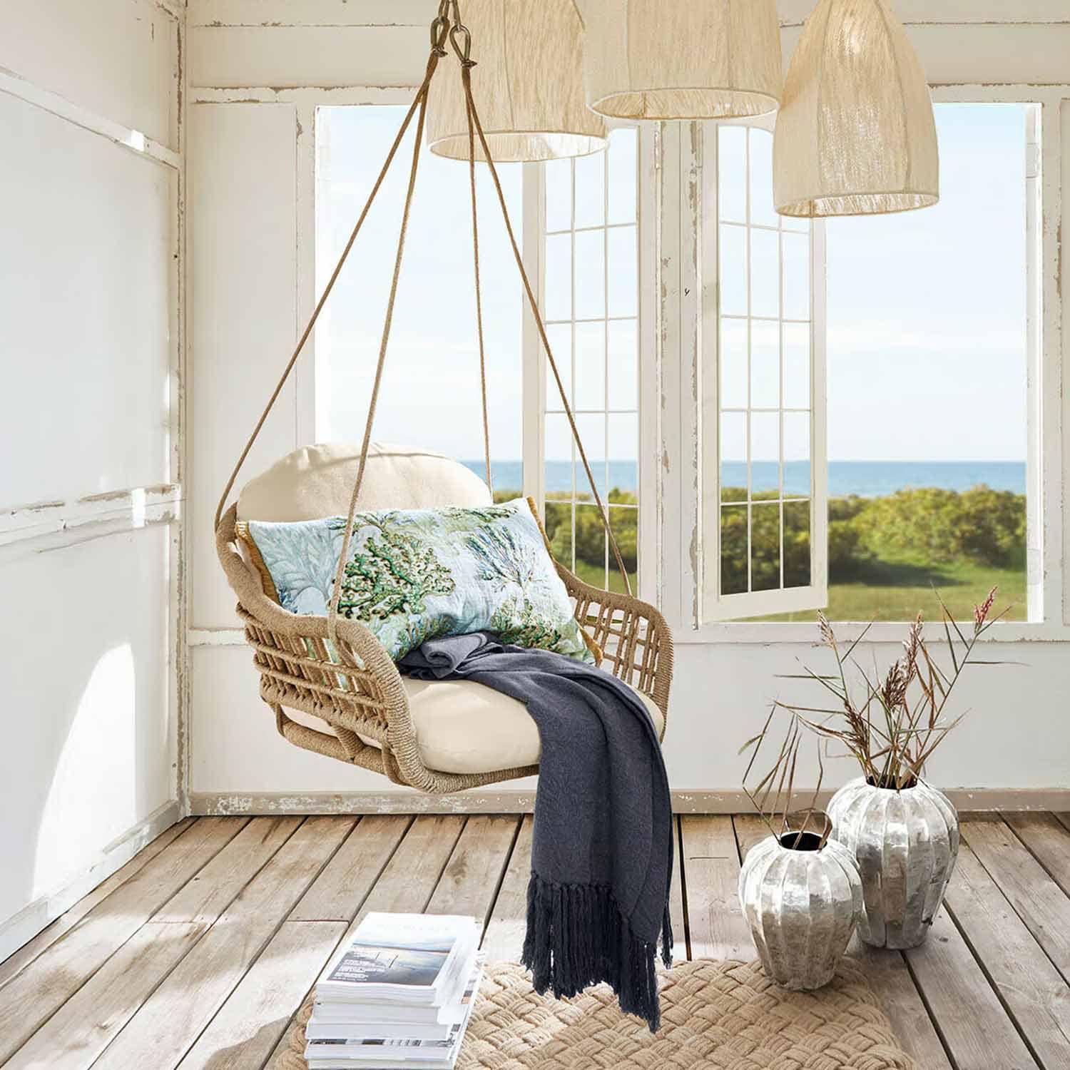 Fauteuil suspendu en rotin avec coussin crème et plaid bleu dans une pièce lumineuse vue mer