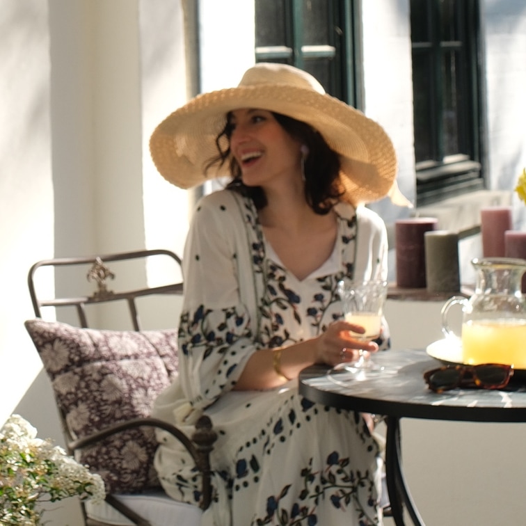 Femme en robe fleurie et chapeau de paille assise à une table avec boisson dans une pièce lumineuse