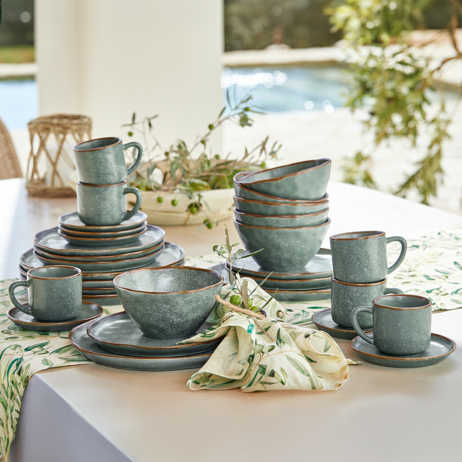 Service de vaisselle en grès vert avec tasses et assiettes sur une table claire décorée de tissu à motifs