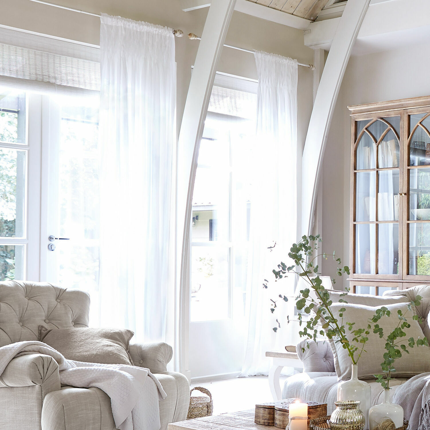 Rideau Otilie Salon lumineux avec fauteuil beige, rideaux blancs et vitrine en bois style vintage