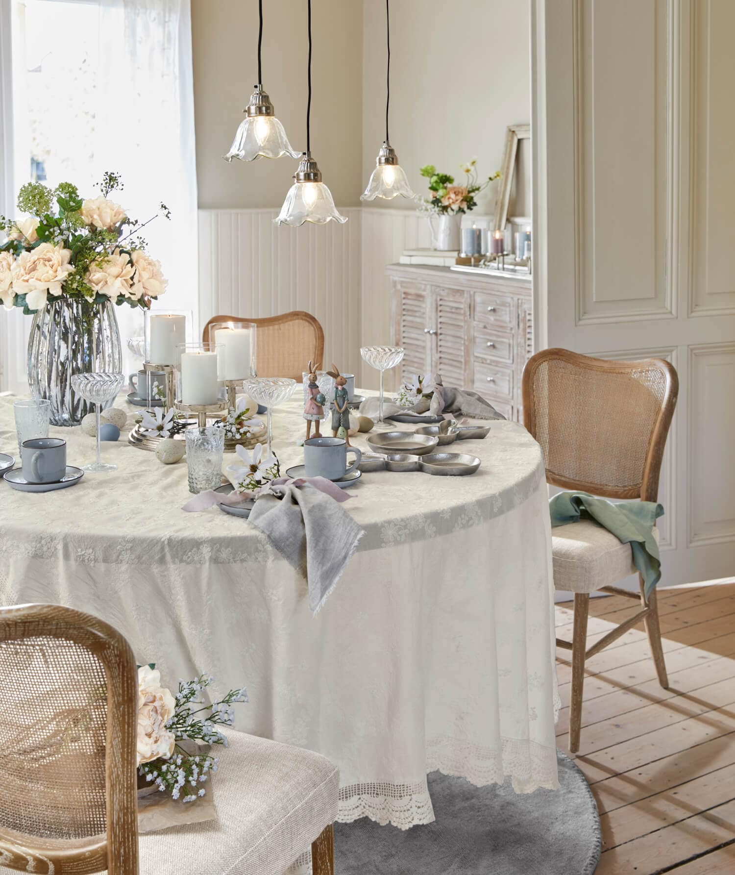 Table claire avec nappe crème et décorations rustiques, chaises en bois et fleurs dans une salle à manger lumineuse