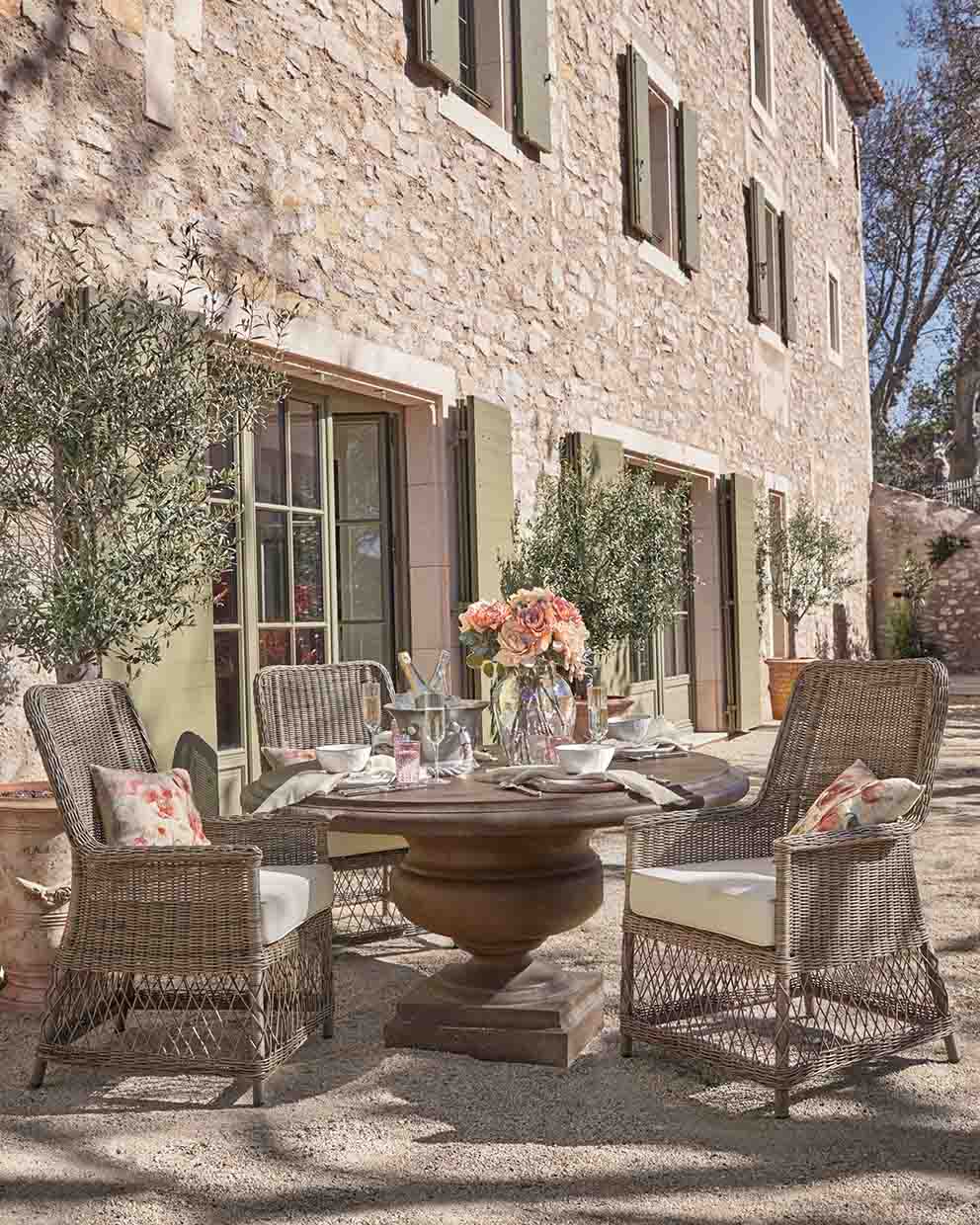 Table ronde en pierre avec quatre chaises en osier et coussins beige dans un jardin ensoleillé