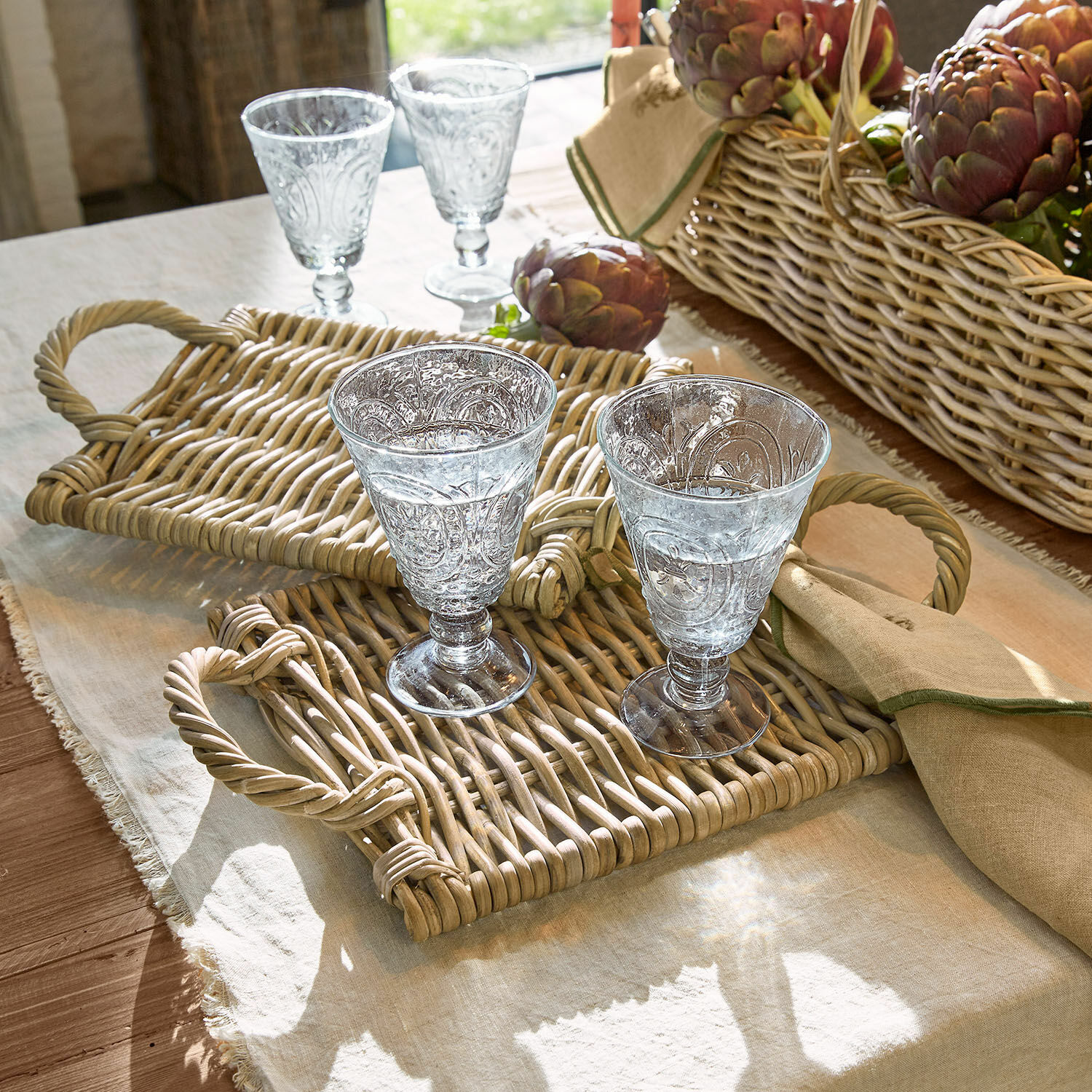 Lot de 2 sets de table Allegretta Deux verres décorés sur plateaux en osier posés sur une nappe en lin avec un panier d'artichauts